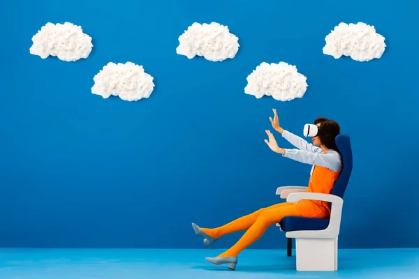 Vista laterale di afro americano in auricolare vr con le mani tese seduto sul sedile su sfondo blu con nuvole — Foto stock