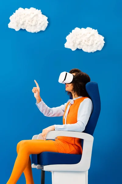 Vista lateral de afroamericano con auriculares vr apuntando con el dedo sobre fondo azul con nubes - foto de stock