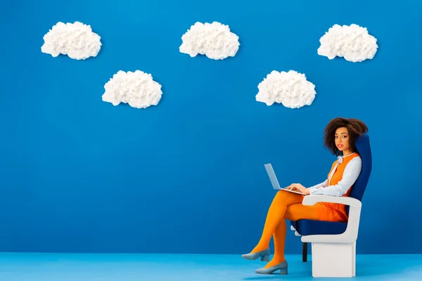 Afro-américaine en robe rétro assise sur un siège et tenant un ordinateur portable sur fond bleu avec des nuages — Photo de stock