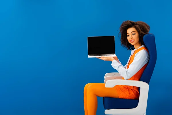 Smiling african american sitting on seat and holding laptop isolated on blue — Stock Photo