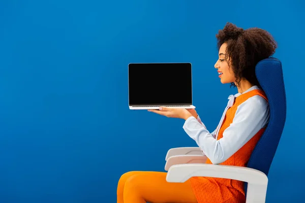 Seitenansicht eines lächelnden Afroamerikaners, der auf seinem Sitz sitzt und Laptop isoliert auf blauem Grund hält — Stockfoto