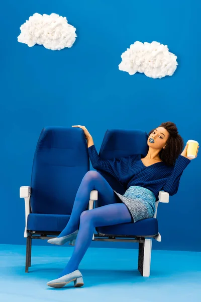 Africano americano sentado en asiento y sosteniendo taza de papel sobre fondo azul con nubes - foto de stock