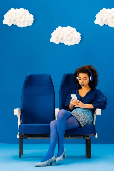 Afroamericano sentado en el asiento, escuchando música y usando un teléfono inteligente sobre fondo azul con nubes - foto de stock