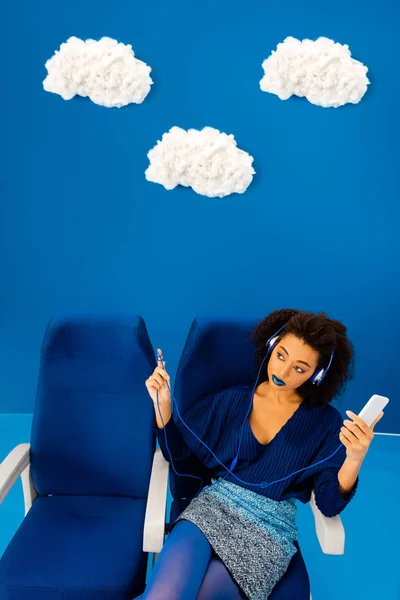 Vue grand angle d'afro-américain assis sur le siège, écoutant de la musique et tenant smartphone sur fond bleu avec des nuages — Photo de stock