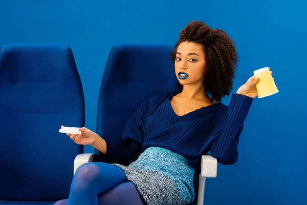 Africano americano sentado no assento e segurando copo de papel isolado em azul — Fotografia de Stock