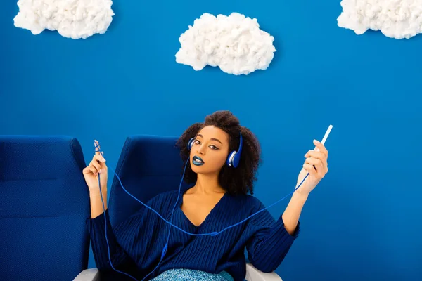 Afro-américain assis sur un siège, écoutant de la musique et utilisant un smartphone sur fond bleu avec des nuages — Photo de stock