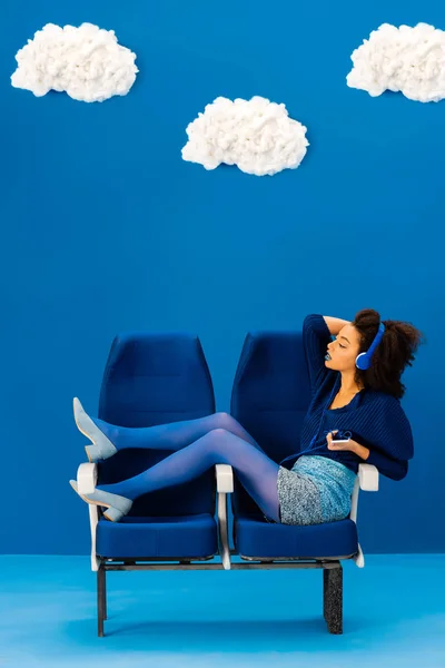 Side view of african american sitting on seats, listening to music on blue background with clouds — Stock Photo