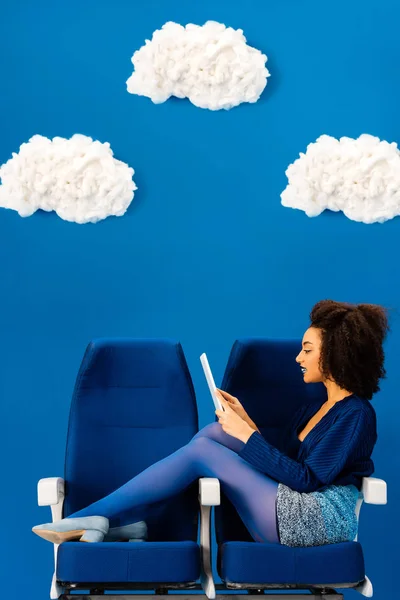 Vista lateral de afroamericano sentado en asientos y usando tableta digital sobre fondo azul con nubes - foto de stock