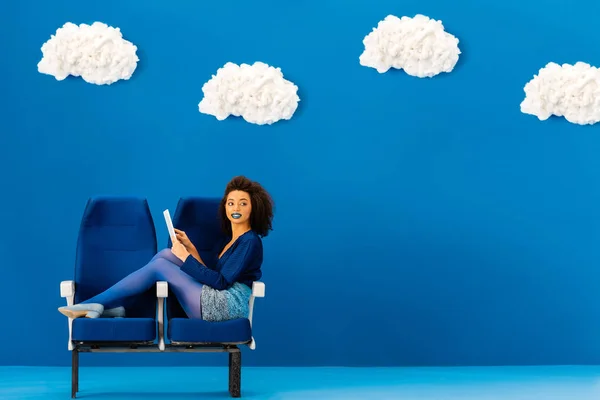 Sonriente afroamericano sentado en asientos y usando tableta digital sobre fondo azul con nubes - foto de stock