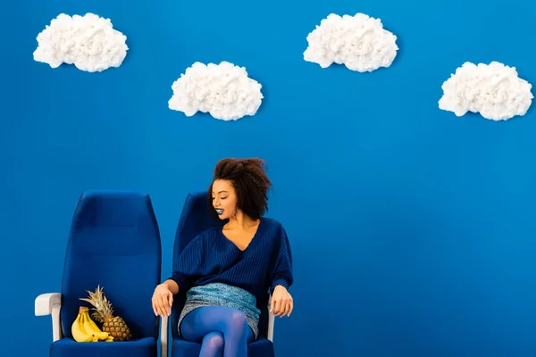 Sonriente afroamericano sentado en el asiento y mirando plátanos y piña sobre fondo azul con nubes - foto de stock