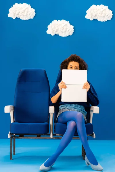 Afro-américain visage obscurcissant avec ordinateur portable et assis sur un siège sur fond bleu avec des nuages — Photo de stock