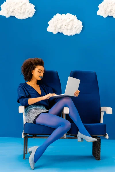 Smiling african american sitting on seat and using laptop on blue background with clouds — Stock Photo