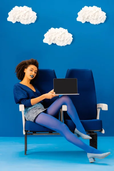 Sorrindo afro-americano sentado no assento e segurando laptop no fundo azul com nuvens — Fotografia de Stock