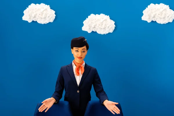 Sonriente azafrán americano azafata apuntando con las manos a los asientos sobre fondo azul con nubes - foto de stock