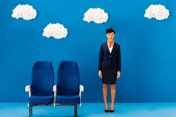 Sonriente asistente de vuelo afroamericano de pie cerca de asiento sobre fondo azul con nubes - foto de stock