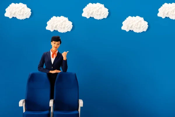 Sorrindo afro-americano comissário de bordo mostrando como perto do assento no fundo azul com nuvens — Fotografia de Stock