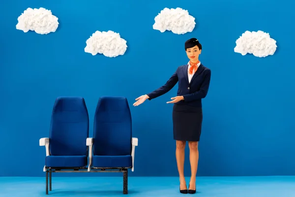 Afro-americano comissário de bordo apontando com as mãos para assentos no fundo azul com nuvens — Fotografia de Stock