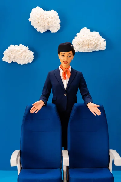 Sorridente assistente di volo afroamericano che punta con le mani ai sedili su sfondo blu con nuvole — Foto stock