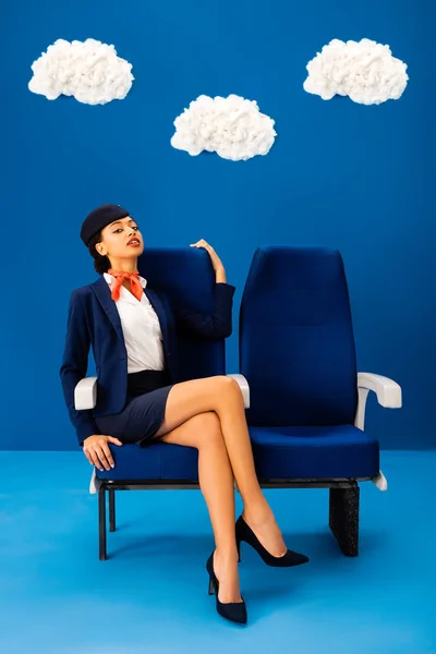 Auxiliar de vuelo afroamericano sentado en asiento sobre fondo azul con nubes - foto de stock