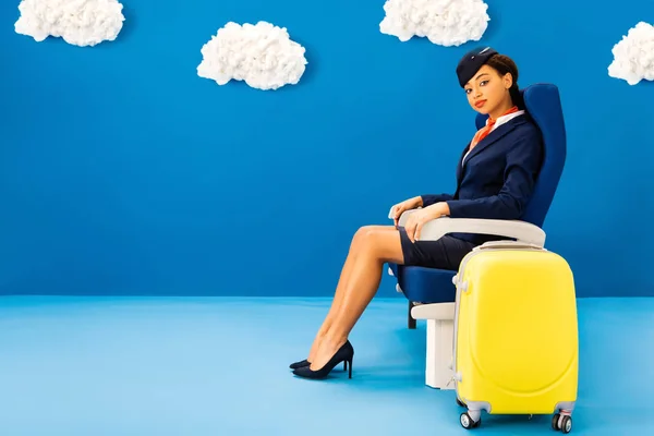 Auxiliar de vuelo afroamericano sentado en el asiento cerca de la bolsa de viaje sobre fondo azul con nubes - foto de stock