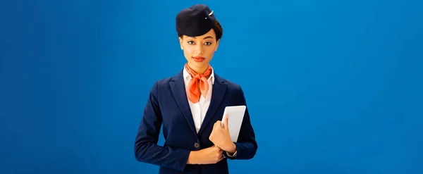 Panoramaaufnahme eines lächelnden afrikanisch-amerikanischen Flugbegleiters, der ein digitales Tablet isoliert auf blauem Grund hält — Stockfoto