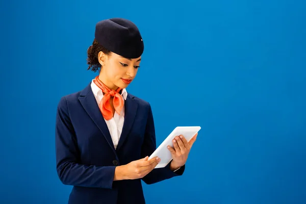 Улыбающаяся африканская стюардесса, использующая цифровой планшет, изолированный на голубом — стоковое фото