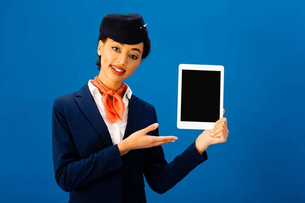 Lächelnder afrikanisch-amerikanischer Flugbegleiter zeigt mit der Hand auf digitales Tablet isoliert auf blauem Grund — Stockfoto