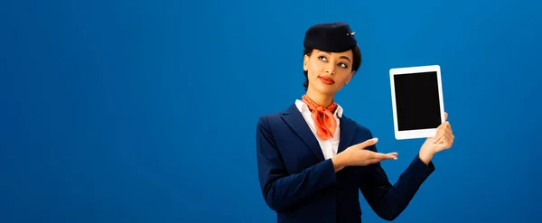 Panoramic shot of smiling african american flight attendant pointing with hand at digital tablet isolated on blue — Stock Photo