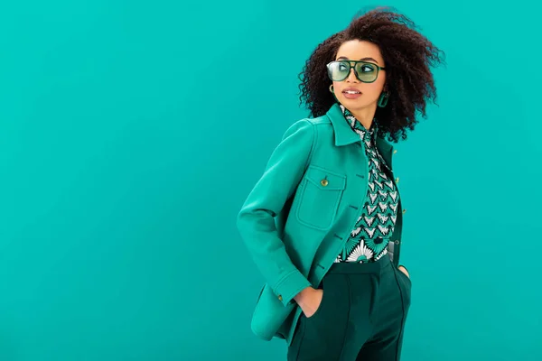Stylish african american woman in jacket with hands in pocket looking away isolated on turquoise — Stock Photo