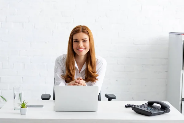 Erfolgreiche Geschäftsfrau lächelt in die Kamera neben Laptop und Handy auf dem Tisch — Stockfoto
