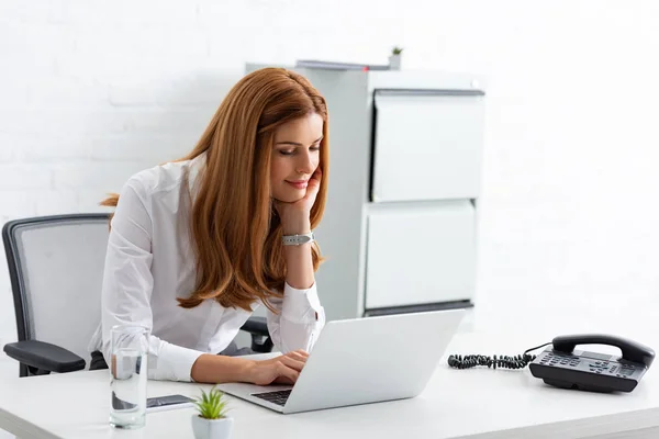 Donna d'affari sorridente utilizzando il computer portatile vicino al telefono sul tavolo — Foto stock