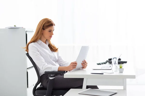 Vista lateral de la mujer de negocios leyendo dossier cerca de gadgets en la mesa - foto de stock