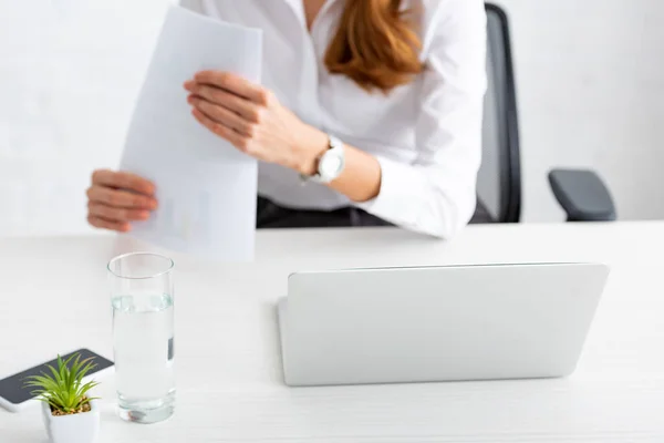 Concentration sélective de l'ordinateur portable et verre d'eau et femme d'affaires tenant des documents à table — Photo de stock