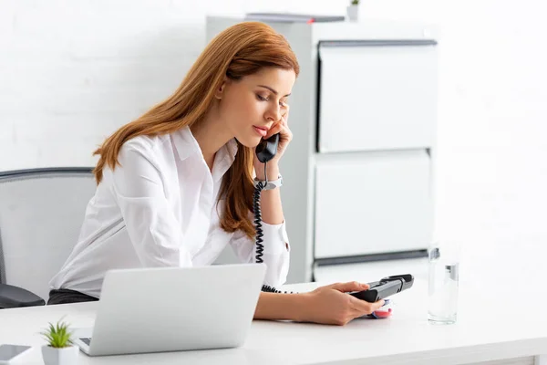 Bella donna d'affari che parla al telefono vicino a gadget e vetro d'acqua sul tavolo — Foto stock