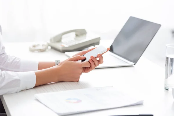 Ausgeschnittene Ansicht einer Geschäftsfrau mit Smartphone in der Nähe von Dokumenten und Laptop auf dem Tisch — Stockfoto
