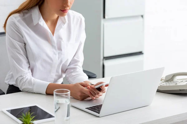 Vista cortada de empresária segurando smartphone perto de laptop e tablet digital na mesa — Stock Photo