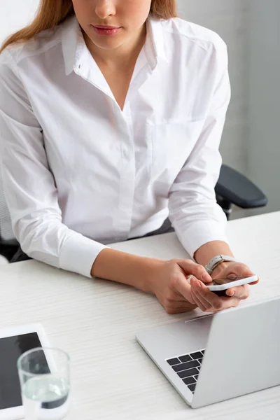 Cropped view of businesswoman holding smartphone near gadgets and glass of water on table — Stock Photo