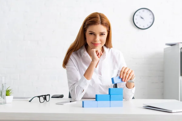 Femme d'affaires réussie souriant à la caméra tout en empilant pyramide de marketing de blocs de construction sur la table — Photo de stock