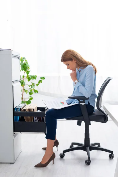 Seitenansicht einer Geschäftsfrau, die Papiere mit Graphik hält, während sie in der Nähe eines offenen Schranktreibers sitzt — Stockfoto