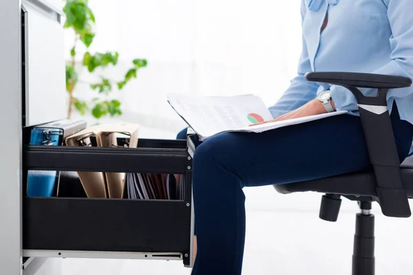 Vista recortada de la mujer de negocios en la silla que sostiene los papeles con el gráfico cerca del conductor abierto del gabinete - foto de stock