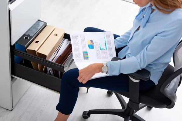 Vista de ángulo alto de la mujer de negocios sosteniendo papeles con gráficos mientras está sentada cerca del conductor del gabinete abierto - foto de stock