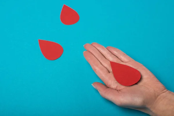 Vista recortada de la palma de la mujer con gotas decorativas de sangre sobre fondo azul, concepto del día mundial de la salud - foto de stock