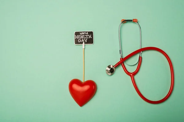 Vista superior del estetoscopio, corazón decorativo y tarjeta con letras del día de la salud mundial sobre fondo verde - foto de stock