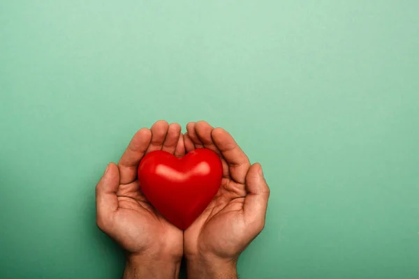 Vista superior del corazón rojo decorativo en las manos del hombre sobre fondo verde, concepto del día mundial de la salud - foto de stock