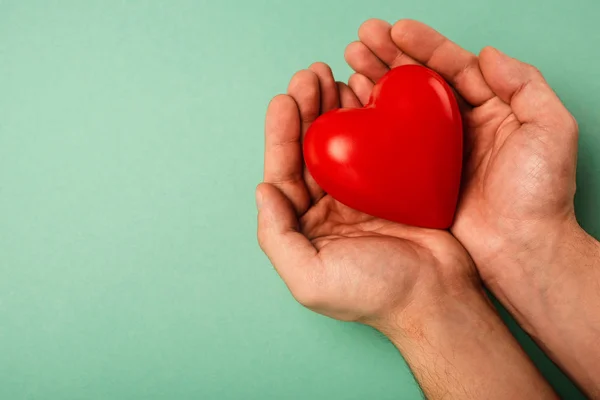 Vista recortada del corazón rojo decorativo en las manos del hombre sobre fondo verde, concepto del día mundial de la salud - foto de stock