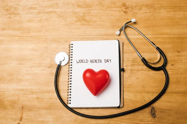 Top view of stethoscope, decorative heart on notebook with world health day lettering on wooden background — Stock Photo