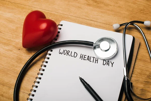 Stethoscope, decorative heart, pen on notebook with world health day lettering on wooden background — Stock Photo