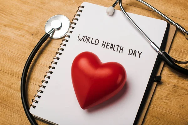 Estetoscopio, corazón rojo decorativo en el cuaderno con letras del día de la salud mundial sobre fondo de madera - foto de stock