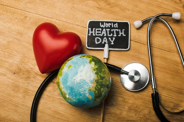 Top view of decorative red heart, stethoscope, globe and card with world health day lettering on wooden background — Stock Photo