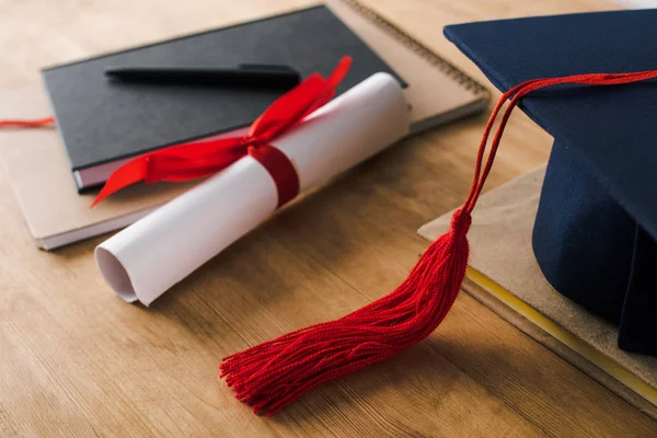 Enfoque selectivo de cuadernos, bolígrafo, diploma y gorra de graduación con borla roja sobre fondo de madera - foto de stock